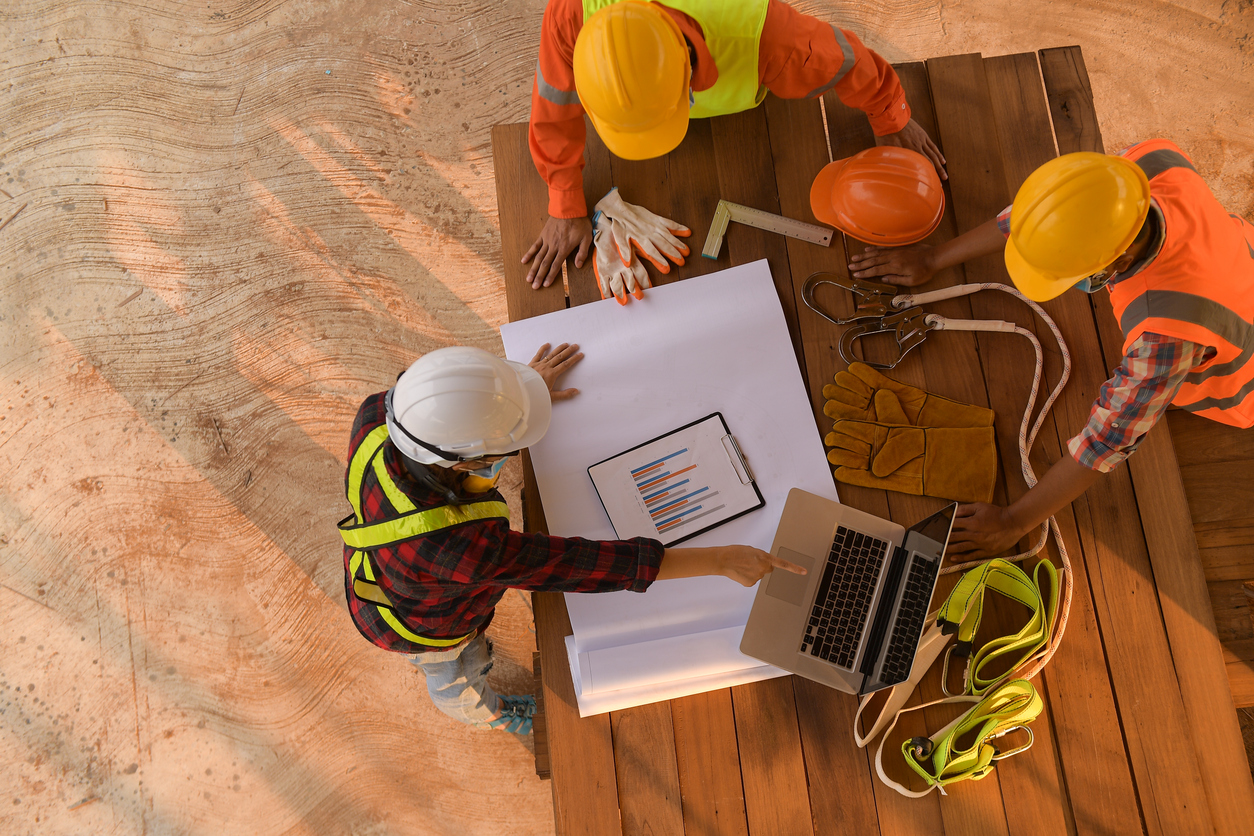 Vista superior de três trabalhadores da construção civil com equipamentos de segurança (capacetes, coletes e arreios) em uma reunião no canteiro de obras, analisando plantas e um laptop sobre uma mesa de madeira