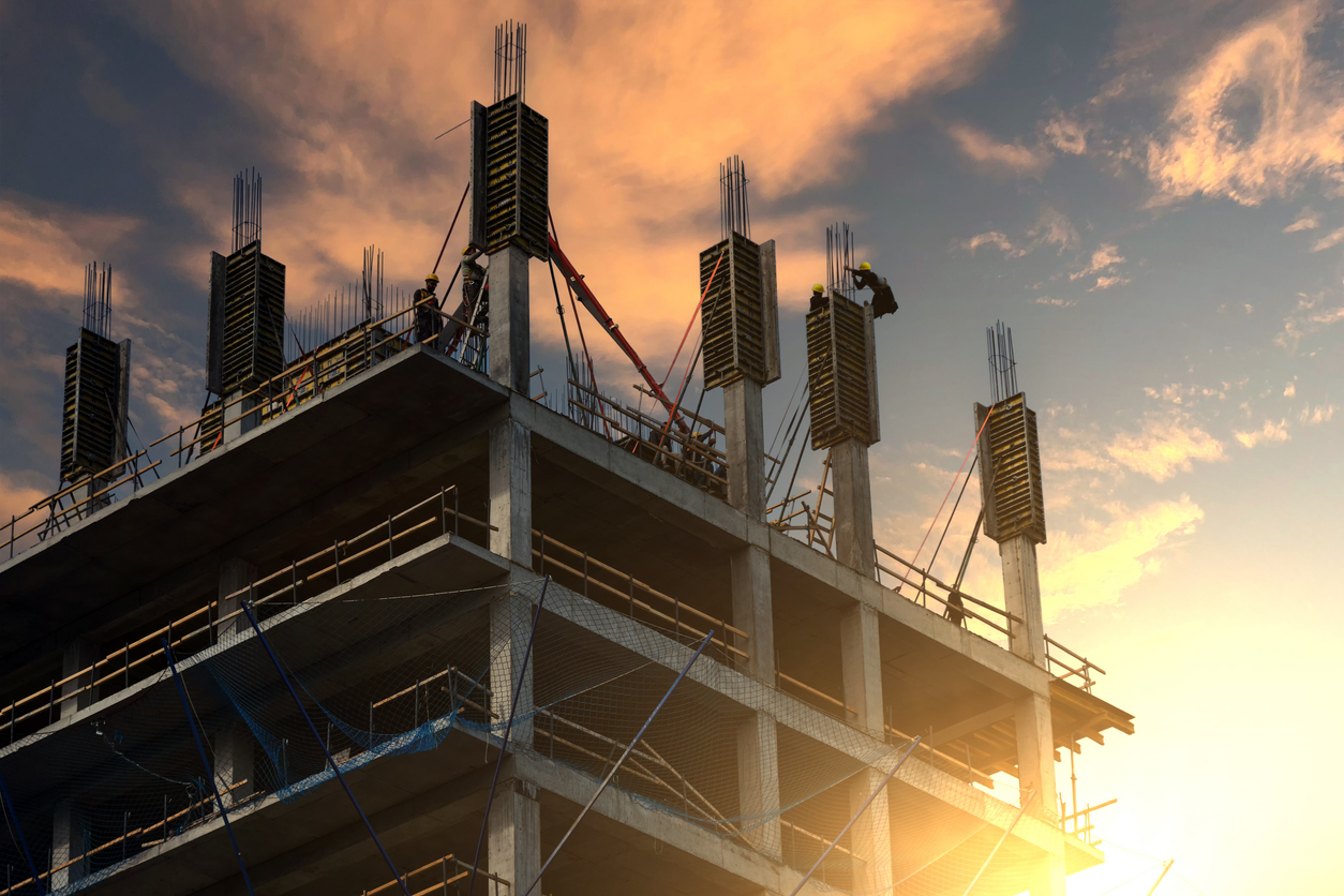 Trabalhadores na cobertura de um edifício em construção de concreto, com o sol baixo ao fundo.