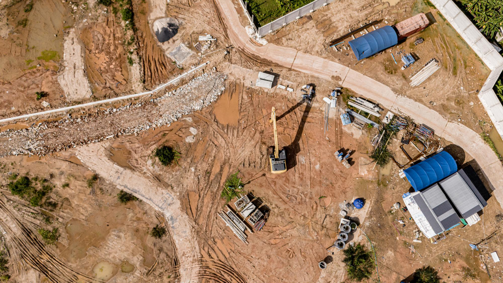 Vista aérea de canteiro de obras com escavadeira, vias de terra, materiais de construção e estruturas provisórias.