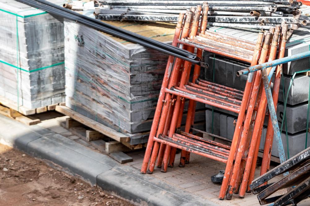 Materiais de construção empilhados em obra, com blocos, paletes e estruturas metálicas apoiadas ao lado.