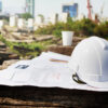 Capacete de segurança sobre plantas técnicas em um canteiro de obras corporativo, com estruturas e prédios ao fundo.