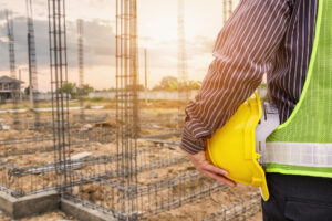 Profissional da construção civil segurando capacete amarelo diante de obra em fase de fundação, com armações de aço e escavações no terreno.