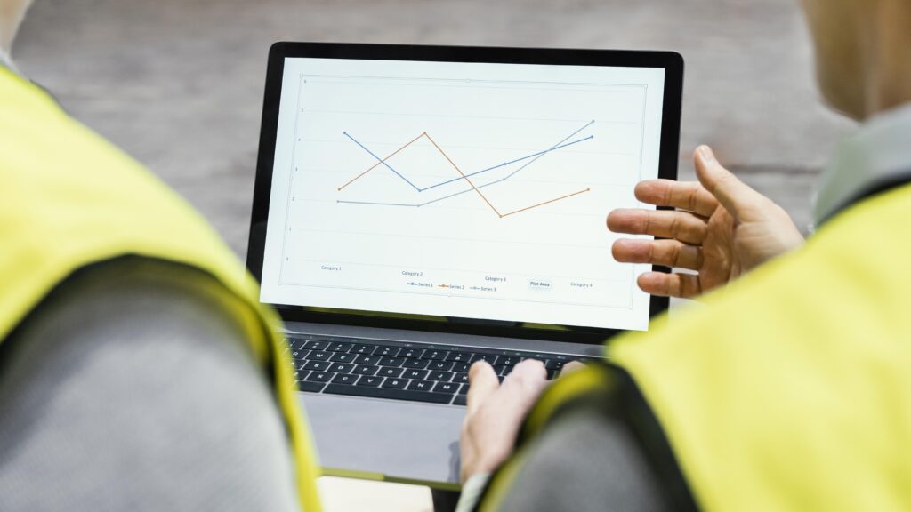 Profissionais com coletes de segurança analisam gráfico de linhas em um notebook.