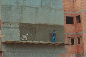 Dois trabalhadores da construção civil aplicam reboco na fachada de um prédio usando andaime de madeira.