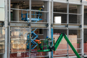 Trabalhadores utilizam plataformas elevatórias para instalar vidros na fachada de um prédio em construção.