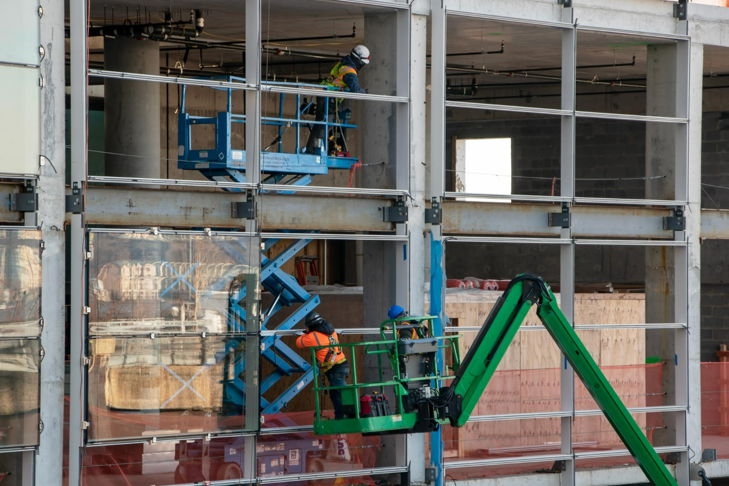Trabalhadores utilizam plataformas elevatórias para instalar vidros na fachada de um prédio em construção.