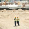 Dois profissionais da construção civil usando capacetes e coletes de segurança conversam em um canteiro de obras amplo, cercado por estruturas em construção e equipamentos ao fundo.