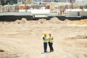 Dois profissionais da construção civil usando capacetes e coletes de segurança conversam em um canteiro de obras amplo, cercado por estruturas em construção e equipamentos ao fundo.