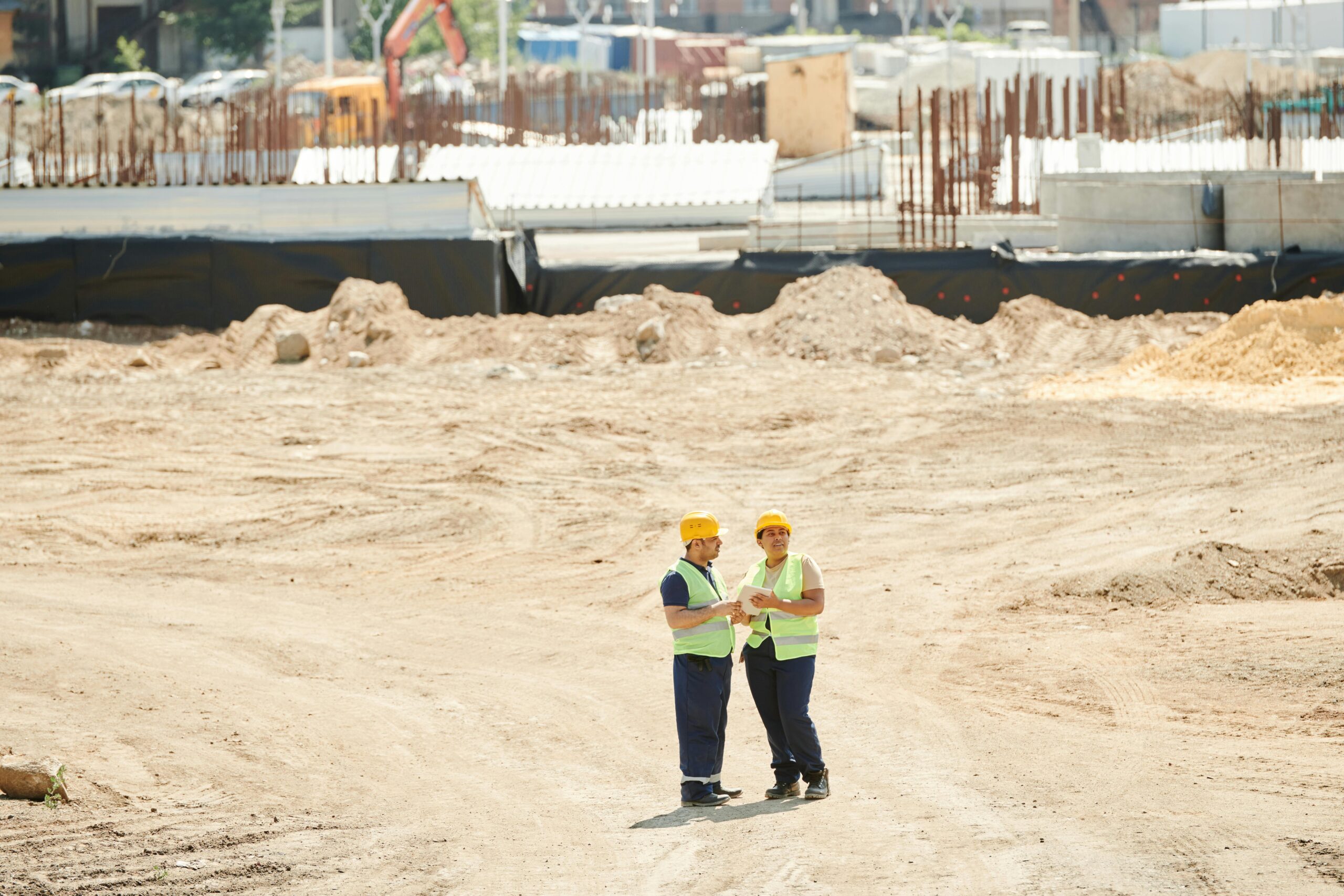 Dois profissionais da construção civil usando capacetes e coletes de segurança conversam em um canteiro de obras amplo, cercado por estruturas em construção e equipamentos ao fundo.