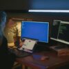 Pessoa trabalhando em um ambiente escuro com dois monitores exibindo códigos e um notebook sobre a mesa iluminada.