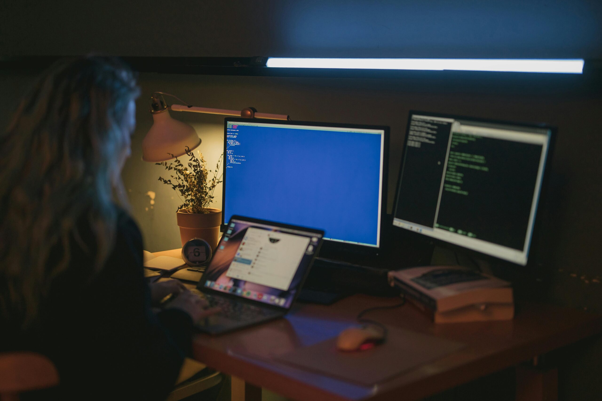 Pessoa trabalhando em um ambiente escuro com dois monitores exibindo códigos e um notebook sobre a mesa iluminada.