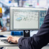 Profissional utilizando computador com software de modelagem ou engenharia industrial, enquanto trabalhador aparece ao fundo em ambiente fabril.