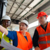 Três profissionais usando capacetes e coletes de segurança inspecionam equipamentos em ambiente industrial.