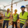 Profissionais da construção civil utilizando capacetes e coletes de segurança em canteiro de obras, observando o andamento de uma construção.