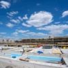Canteiro de obras com estruturas em construção, fundações aparentes, materiais de engenharia e andaimes sob céu azul com nuvens.