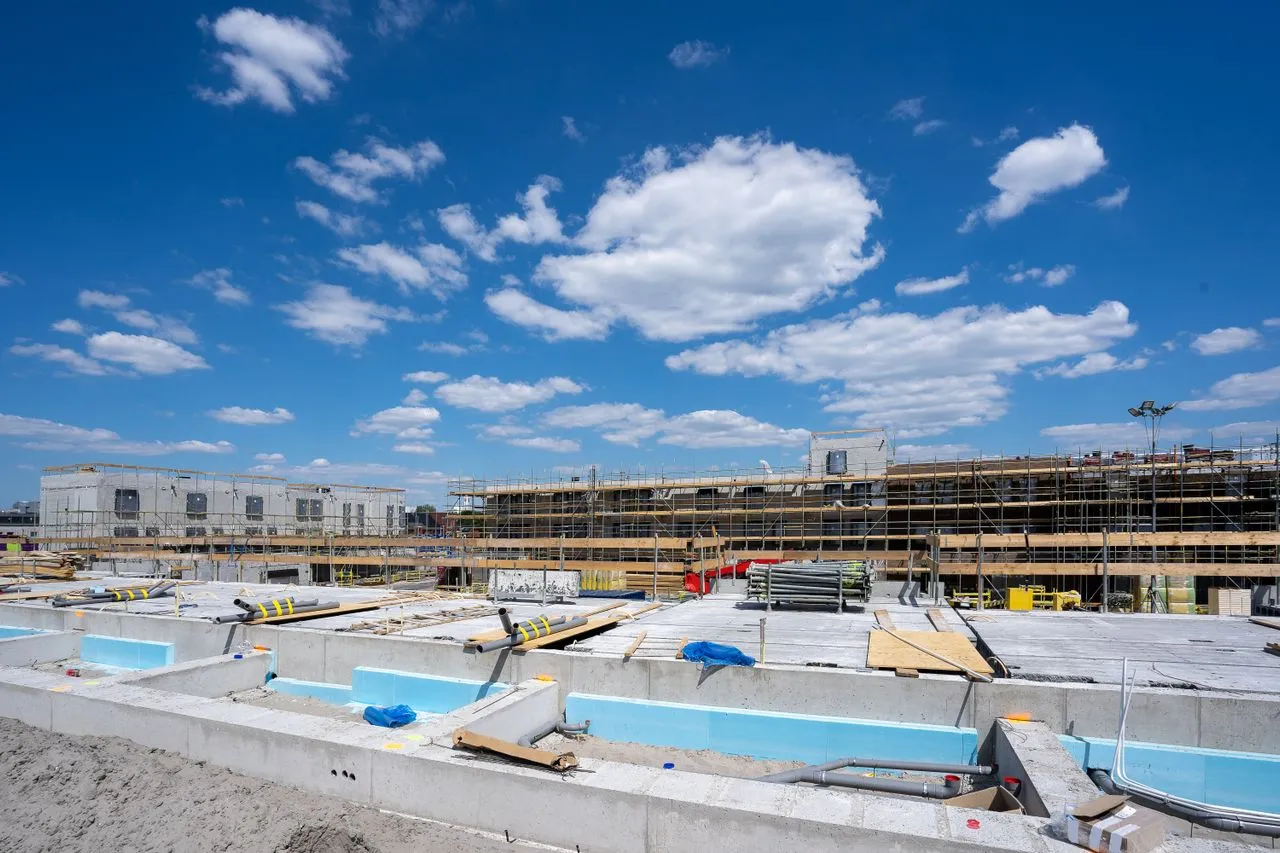 Canteiro de obras com estruturas em construção, fundações aparentes, materiais de engenharia e andaimes sob céu azul com nuvens.