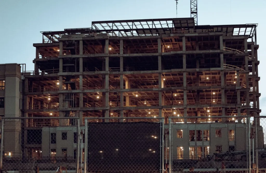 Prédio de múltiplos andares em construção, com estrutura de concreto aparente e iluminação interna acesa ao entardecer.